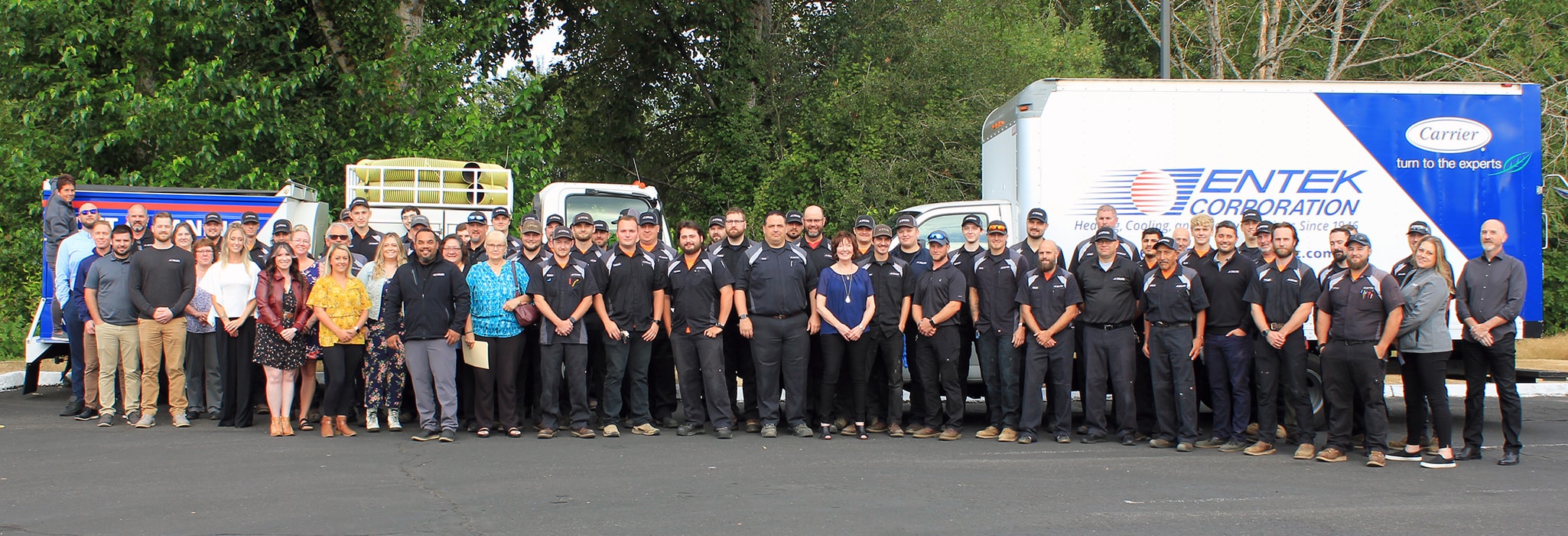 The Entek Corporation team outside lined up outside in front of their service trucks with large trees with green leaves behind them in the background.