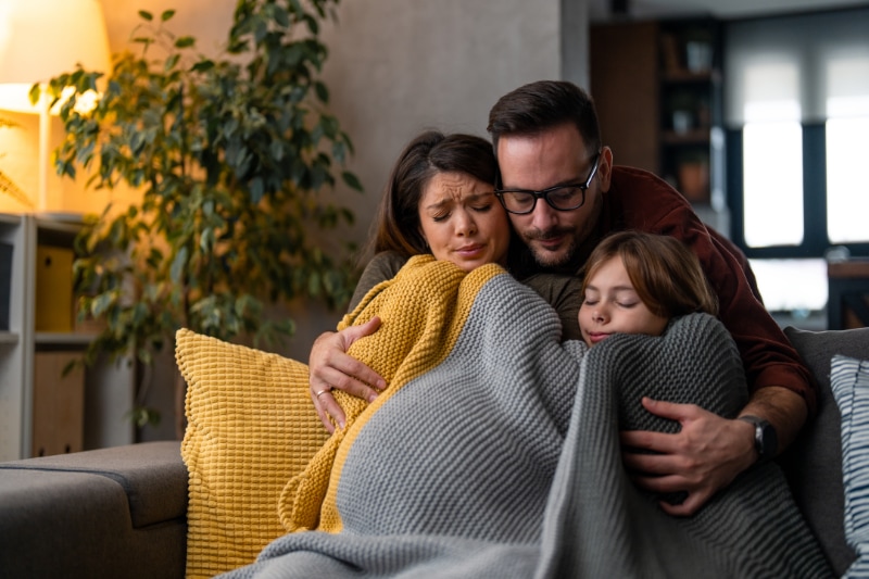 Why Isn’t My Furnace Heating My Entire Home? Image is a photograph of a family of 3 snuggling on the couch in a cozy blanket.