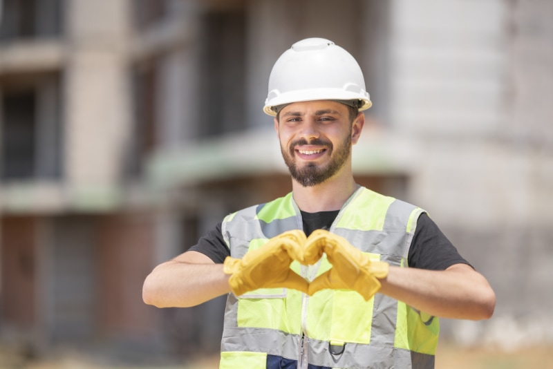 How to Join the HVAC Industry. Image is a photograph of an HVAC professional wearing a white helmet and reflective vest working outside. He is making a heart with his hands.