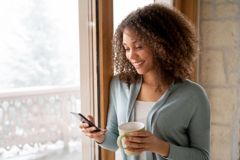 Why Choose a Variable-Speed Heat Pump? Image is a photograph of a woman texting by a window and holding a coffee mug.